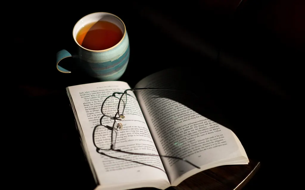 An open book, reading glasses and a cup of tea on a dark table